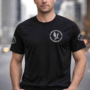 May include: Black crew neck t-shirt with white text and a logo for the Los Angeles Police Department Special Weapons and Tactics. The shirt has short sleeves and is worn with black trousers. The image is taken outdoors.