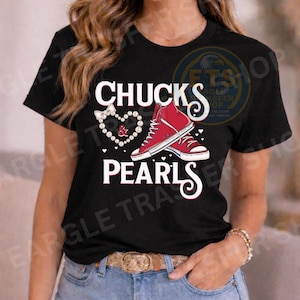 May include: Black t-shirt with the words "CHUCKS PEARLS" in white script. The design includes a pair of red high-top sneakers and a pearl necklace in the shape of a heart. The shirt is worn with blue jeans and a brown belt.