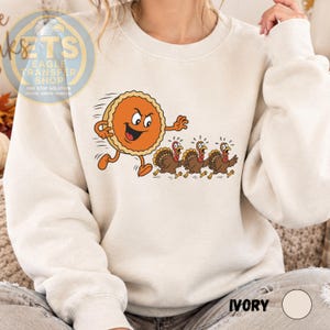 May include: An ivory crewneck sweatshirt featuring a graphic print of a cartoon pumpkin pie with arms and legs chasing three brown turkeys. The pie has a mischievous expression, while the turkeys appear to be running away in a state of panic.