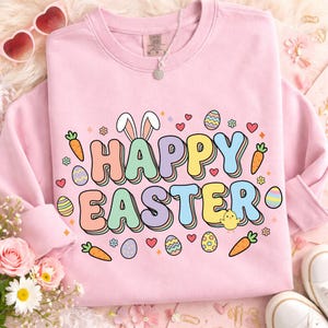 May include: A pale pink sweatshirt featuring the text "HAPPY EASTER" in a colourful, bubble-style font. The design incorporates bunny ears, Easter eggs, carrots, and hearts. A necklace and sunglasses are visible in the background.