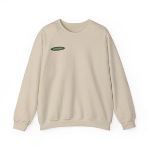 Puede incluir: Sudadera color crema con mangas largas y cuello redondo. Un pequeño parche ovalado verde en el pecho presenta la palabra "reminder!" en blanco y minúsculas. La sudadera es de material suave, adecuada para uso casual.