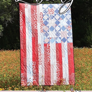 May include: A patriotic quilt with a design resembling the American flag. The quilt features red and white stripes, a blue square with white stars, and the word "Liberty" in script. The quilt is displayed outdoors.