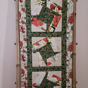 May include: A green, red, and white Christmas table runner with a geometric pattern of poinsettias, holly, and a floral design. The runner is hanging on a bamboo rack.