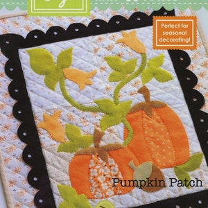 May include: A quilted fabric square with a pumpkin patch design. The design features orange pumpkins, green leaves, and yellow flowers on a white background with a floral pattern. The square is framed with a black scalloped border. Text includes "Figs and Woolies" and "Pumpkin Patch".