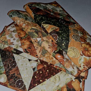 May include: A folded, quilted table runner with a patchwork design. The fabric features a fall harvest theme with pumpkins, leaves, and orange slices in shades of orange, brown, and cream. The runner is made of multiple fabric pieces sewn together.