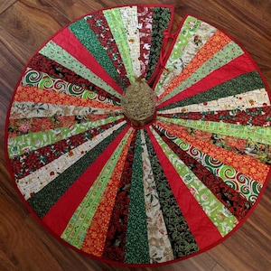 May include: A red, green, and white quilted Christmas tree skirt with a variety of holiday-themed patterns. The skirt is made of fabric scraps and has a red binding around the edge.