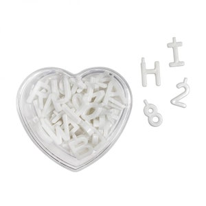 May include: A clear, heart-shaped container filled with white alphabet letters. Several individual white letters and numbers, including the number 2, are scattered nearby. The letters are in a sans-serif font.
