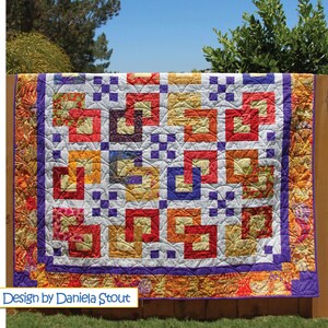 May include: A colorful quilt with a geometric pattern of squares and rectangles in shades of red, orange, yellow, and purple. The quilt is displayed outdoors, draped over a wooden fence. The text "Design by Daniela Stout" is visible.