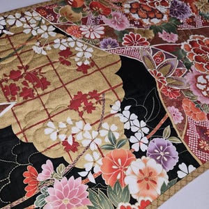 May include: Quilted table runner with a Japanese floral design. The runner features a gold, black, and red color scheme with intricate floral patterns. The edges are trimmed with a gold border, adding a touch of elegance to the home decor item.