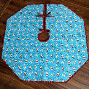 May include: A blue and red quilted Christmas tree skirt with a Santa and reindeer pattern. The skirt has a hole in the center for the tree trunk.