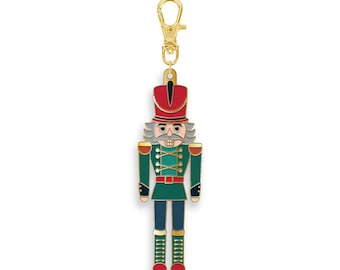 May include: A gold-toned metal keychain featuring a nutcracker charm. The nutcracker is decorated with red, green, and dark blue enamel, accented with gold-colored details on the uniform, hat, and boots. It includes a gold lobster claw clasp.