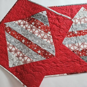 May include: Red and gray quilted table runner with a diamond pattern featuring snowflakes and stars. The runner has a quilted design with a red background and a diamond pattern in the center. The diamond pattern is made up of gray and white fabric with snowflakes and stars.