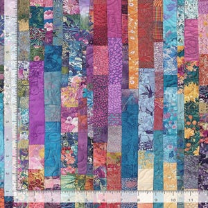 May include: A colorful patchwork quilt with vertical strips of various patterned fabrics. The quilt features a mix of floral, abstract, and textured designs in shades of purple, blue, orange, and pink. A ruler is visible along the left edge.