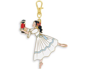 May include: A gold-toned metal keychain charm featuring an enamel illustration of a dancer in a white dress with light blue trim, holding a small red and black nutcracker figure. The dancer has dark hair with a red bow and wears pink ballet slippers.