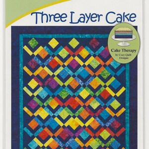 May include: A package for a quilt pattern titled "Three Layer Cake" by Cozy Quilt Designs. The quilt design features a colorful geometric pattern with squares in shades of blue, green, yellow, orange, and purple. The pattern uses 10-inch squares.