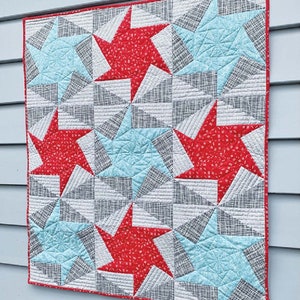 May include: A quilted wall hanging featuring a repeating star pattern. The stars are red and light blue, set against a gray and white background. The quilt is bordered with a red binding and has a geometric design.