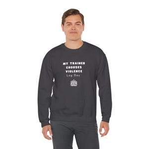 Könnte beinhalten: Dunkelgraues Sweatshirt mit dem weißen Text "MY TRAINER CHOOSES VIOLENCE Leg Day". Das Sweatshirt hat einen Rundhalsausschnitt und lange Ärmel. Unter dem Text befindet sich ein kleines Emblem.