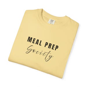 May include: A folded, pale yellow t-shirt with the words "MEAL PREP Society" printed in black. The text is in a bold, sans-serif font and a cursive font. The shirt has a crew neck and a label.