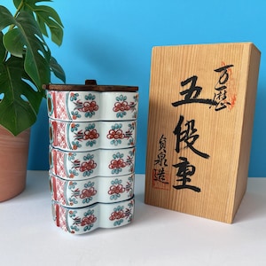 Könnte beinhalten: Ein stapelbarer, fünflagiger japanischer Keramik-Lebensmittelbehälter mit Holzdeckel. Die weißen Keramikbehälter sind mit roten und grünen Blumenmustern verziert. Rechts befindet sich eine Holzkiste mit japanischer Kalligraphie.