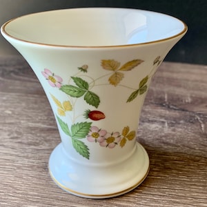 May include: White ceramic vase with a flared top and a pedestal base. The vase is decorated with a strawberry plant design, featuring pink flowers, green leaves, and red strawberries. Gold trim accents the rim and base.