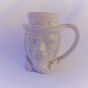 May include: A ceramic mug shaped like Uncle Sam, with a white finish. The mug features a detailed face, a top hat with three stars, and a curved handle. The mug is designed for beverages.