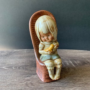 May include: A ceramic figurine of a seated child holding a yellow bird. The child has blonde hair and is wearing a blue skirt. The figurine is seated in a brown wicker chair. The overall color palette is muted earth tones.