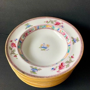 May include: A stack of white porcelain plates with a floral design. Each plate features a pink and gold rim, a central floral motif, and a colorful border with flowers and leaves. The plates are stacked on top of each other.