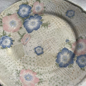 May include: A ceramic plate with a floral design. The plate features a cream-colored background with a pattern of blue and pink flowers. The flowers are surrounded by a subtle, textured pattern. The plate has a slightly scalloped edge.