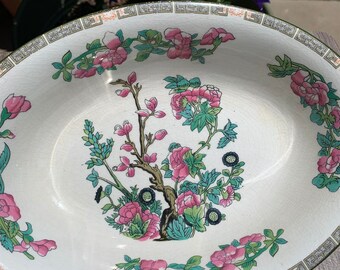 May include: Oval ceramic serving dish with a floral design. The dish features a white background with pink flowers, green leaves, and a brown tree branch. The rim has a decorative border with a green and gold trim.