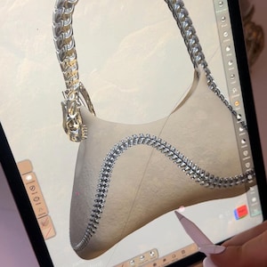 Puede incluir: Representación digital de un bolso beige con asa y ribete de cadena plateada. El bolso se muestra en la pantalla de una tableta, con un lápiz óptico sobre el diseño. El asa presenta un diseño ornamentado.