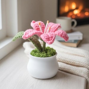 May include: A handmade crochet lily flower in a white ceramic pot. The pink flower has detailed petals and yellow stamens. The pot is filled with green moss. The flower is displayed on a white surface near a window.
