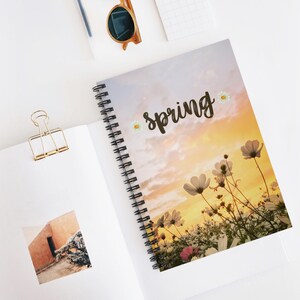 Puede incluir: Cuaderno de espiral con un diseño floral y la palabra "spring" en escritura cursiva. La portada presenta un cielo de puesta de sol y flores de cosmos blancas. Se ve un clip de papel dorado y gafas de sol.