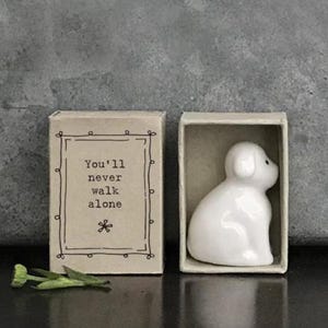 May include: A ceramic white puppy figurine inside a matchbox. The matchbox next to it has the text "You'll never walk alone" in a decorative frame. A small green plant is next to the matchbox.