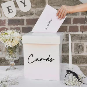 Può includere: Una scatola quadrata bianca per biglietti con la parola Cards stampata in corsivo nero sul davanti. Una busta bianca con la scritta The Buckleys in corsivo nero viene inserita nella fessura superiore. Un vaso di vetro con fiori bianchi e una candela bianca sono vicini.