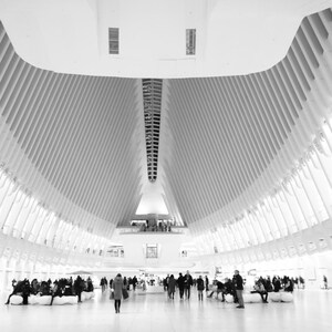 May include: Black and white photograph of a large, modern interior space with a high, curved ceiling. The ceiling is supported by numerous white beams. People are walking and sitting in the open area. The overall design is minimalist and architectural.