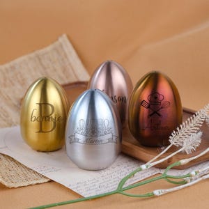 May include: Four egg-shaped metal containers with smooth finishes in gold, silver, rose gold, and a bronze-toned gradient. Each container features custom laser-engraved designs, including names like Bonnie, Martin, and Arthur, and kitchen icons.