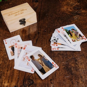 May include: A wooden box with the inscription "William & Mary 21.3.23" sits next to a deck of playing cards. The cards feature photographs and are spread out on a dark wooden surface. Green leaves are in the upper left corner.