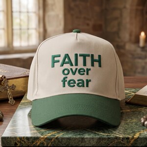 May include: A beige baseball cap with a dark green brim and a dark green embroidered text on the front that reads FAITH over fear. The cap features a structured crown with a small button on top and visible stitching along the brim.
