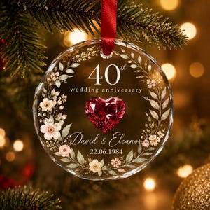 May include: A circular glass ornament hanging from a red ribbon, featuring a floral wreath design, a central red heart-shaped gem, and text reading 40th wedding anniversary, David & Eleanor, and the date 22.06.1984 printed on the clear surface.