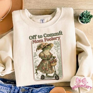 Peut inclure: Sweat-shirt crème avec le texte "Off to Commit Mom Foolery". Le graphisme représente une grenouille en robe et chapeau fleuris sur un scooter. La grenouille porte un panier de fleurs.