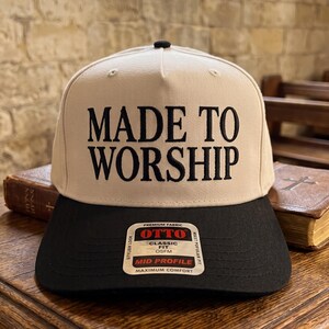 May include: A beige baseball cap with a black brim and a black button on top. The front panel features the embroidered black text MADE TO WORSHIP. A rectangular sticker on the brim displays the text OTTO PREMIUM FABRIC CLASSIC FIT OSFM.