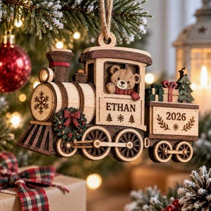 May include: A wooden train Christmas ornament featuring a teddy bear in the cabin, a wreath on the front, and a cargo car with gifts and a tree. The train is engraved with the name ETHAN and the year 2026, accented with snowflake and tree designs.
