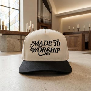 May include: A cream-colored baseball cap with a black brim and a black embroidered script font on the front panel that reads "MADE TO WORSHIP". The cap features a structured crown and a button on the top center, showcasing a two-tone design.