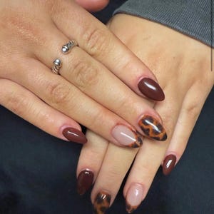 May include: A close-up of a manicure featuring almond-shaped nails. Some nails are painted a deep brown, while others have a clear base with a tortoiseshell design. A silver ring with two small spheres is on one finger.