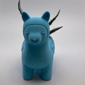 May include: A light blue llama-shaped planter with a small succulent plant. The planter has a smooth finish and features details like ears, eyes, and wings. The plant has green leaves with spiky edges.
