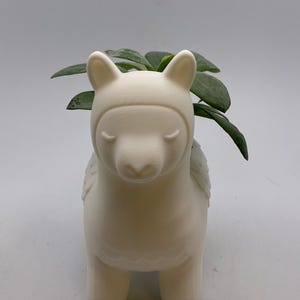 May include: A white ceramic llama planter with a green plant. The llama has closed eyes, a hood, and small wings. The planter is a decorative item for home decor, perfect for adding a touch of whimsy.