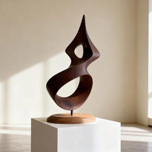 May include: A dark brown, abstract wooden sculpture with a flowing, curved design. The sculpture has a pointed top and a circular opening. It is mounted on a wooden base and a white pedestal.