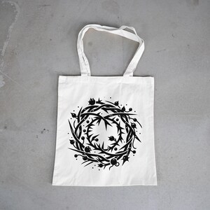 May include: A white tote bag featuring a centered black graphic print of a circular crown of thorns. The design consists of intertwined sharp branches, spikes, and small round berries arranged in a wreath pattern with two handles at the top.
