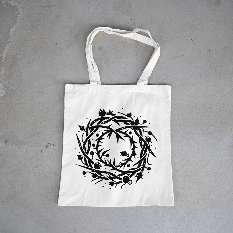 May include: A white tote bag featuring a centered black graphic print of a circular crown of thorns. The design consists of intertwined sharp branches, spikes, and small round berries arranged in a wreath pattern with two handles at the top.