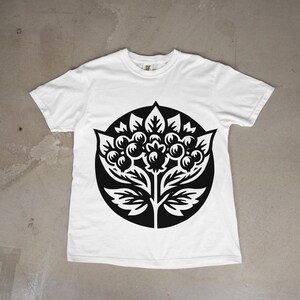 May include: A white short-sleeve t-shirt featuring a large, circular black graphic print on the chest. The design depicts a stylized botanical illustration of a plant with rounded berries, pointed leaves, and serrated foliage elements.